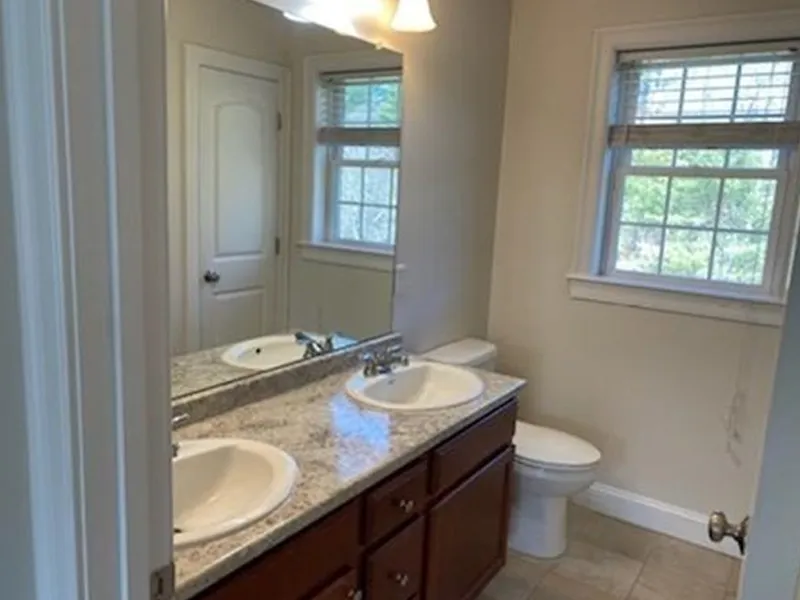 Bathroom with double sinks, granite countertop, toilet, and two windows with blinds.