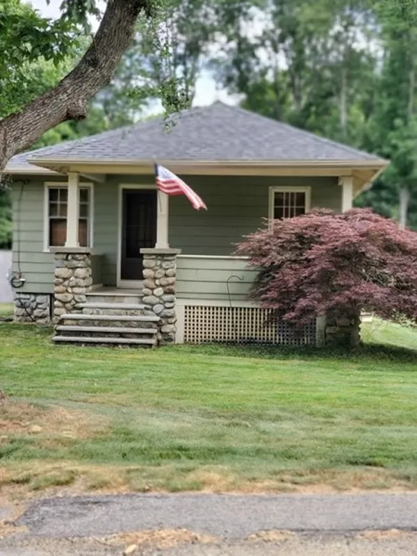Small green house with porch, stone pillars, an American flag, and a red-leaf bush.