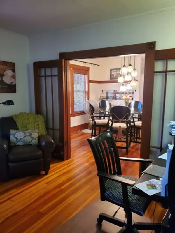 A home office with a desk and chair, opening to a dining area with wooden floors.
