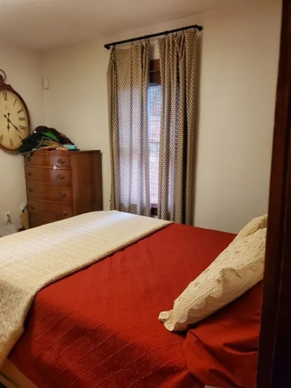Small bedroom with a red bedspread, dresser, large wall clock, and curtained window.