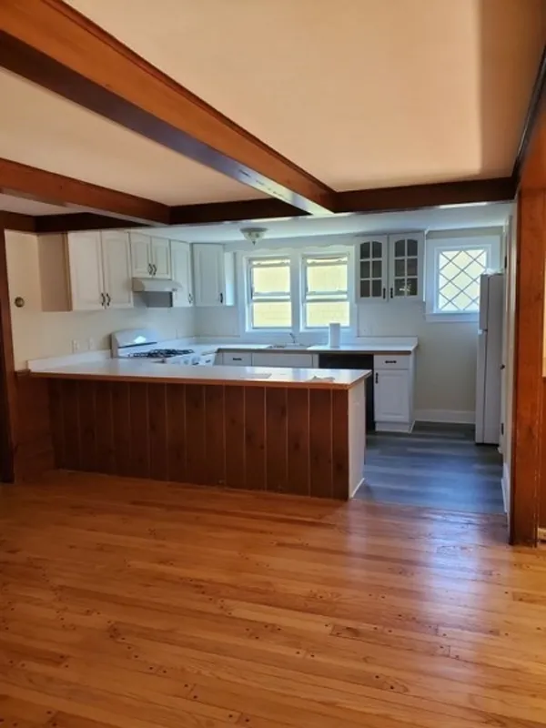 A kitchen with white cabinets, wood beams, and a wooden floor open to a living area.