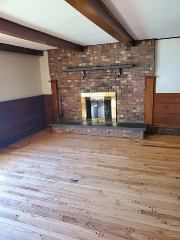 Empty room with wood floors, a brick fireplace, and exposed wooden ceiling beams.