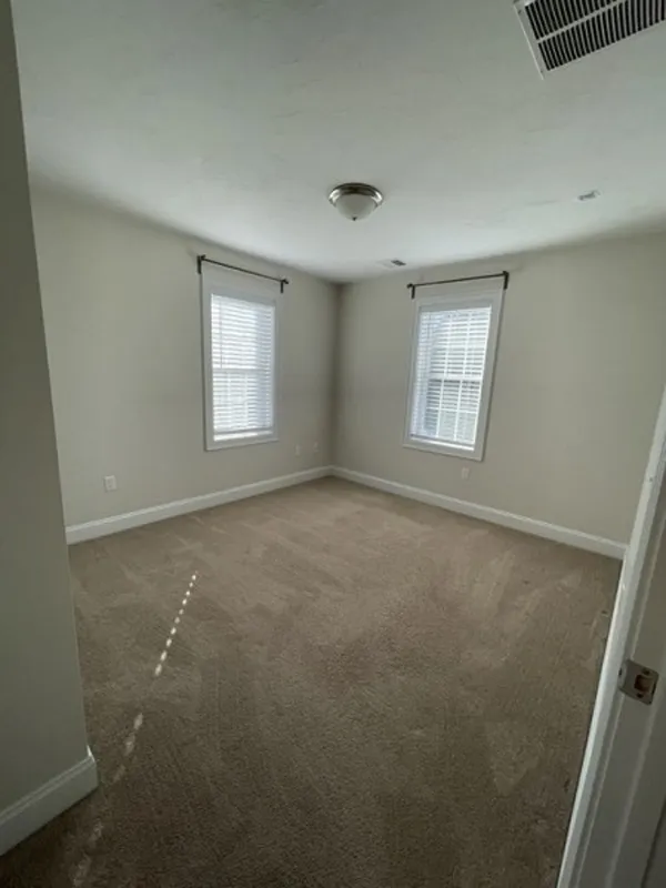 Empty carpeted room with two windows, beige walls, and a ceiling light fixture.