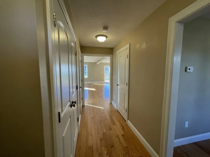 Hallway with wooden floors, beige walls, white doors, and sunlight in the adjoining room.