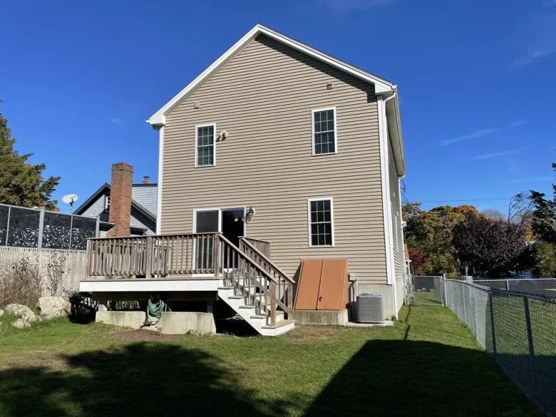 Two-story tan house with a small deck, stairs, and a fenced backyard on a sunny day.