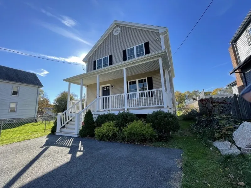 Two-story beige house with front porch, white railing, and driveway on a sunny day.