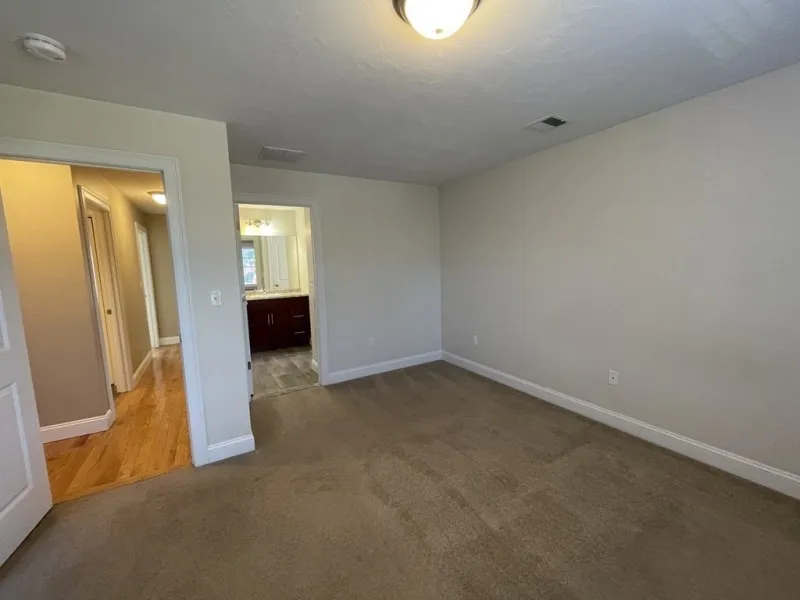 Empty carpeted bedroom with beige walls, a ceiling light, and a view into a bathroom.