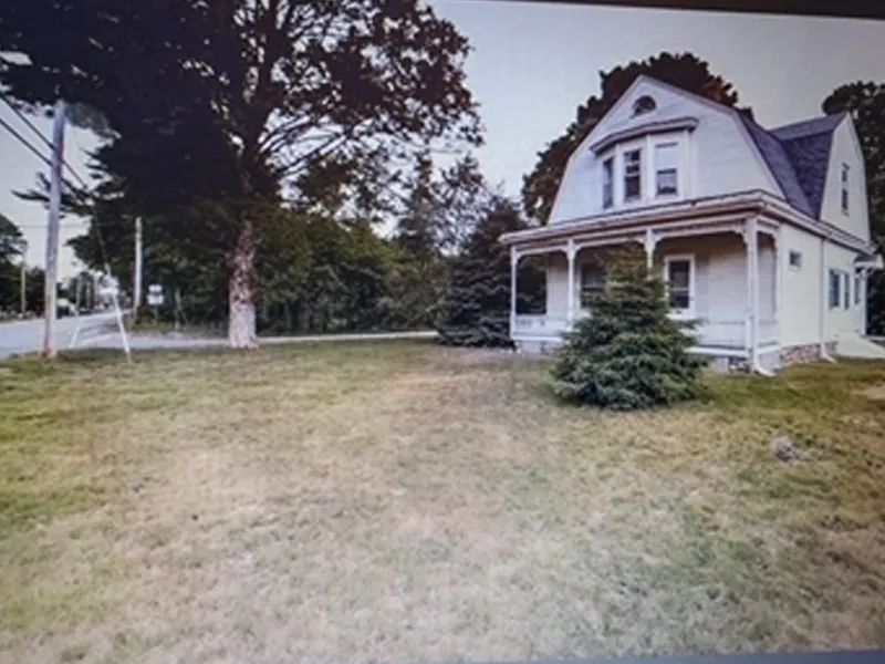 A beige house with a porch and trees in the yard beside a road.