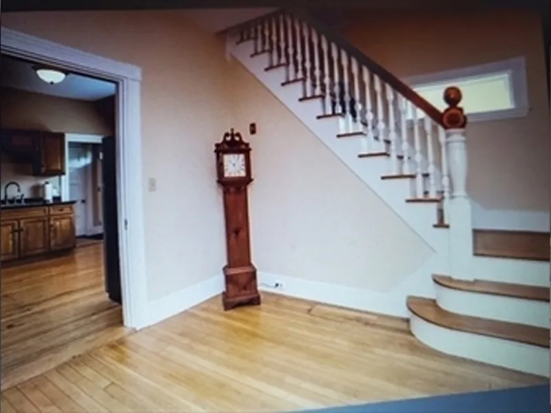 A wooden grandfather clock stands by a staircase in a room with hardwood floors.