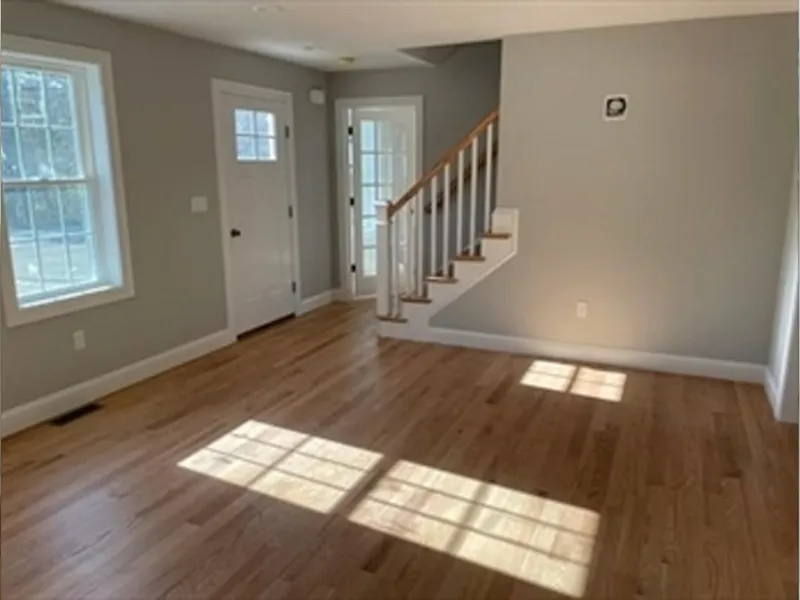 Sunlit empty room with hardwood floors, gray walls, windows, and a staircase.