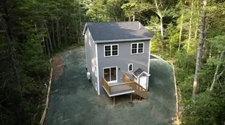 Two-story gray house with small porch surrounded by trees and gravel driveway.