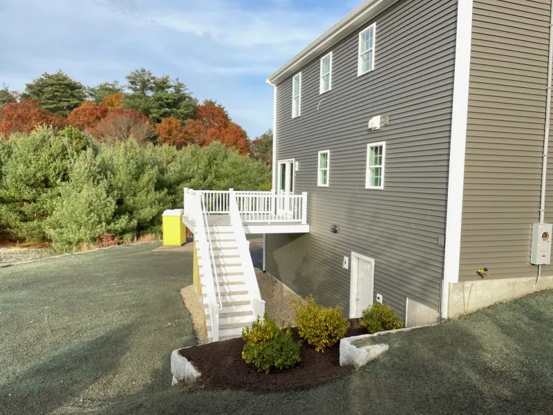Gray house with white stairs and deck, surrounded by trees and a sloped yard.