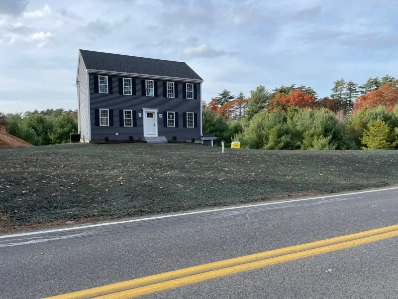 Two-story gray house on a grassy hill beside a road, with trees in the background.