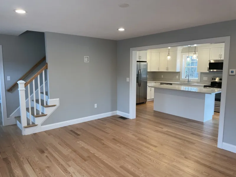 Open modern kitchen with white cabinets and wood floors next to stairs and living area.