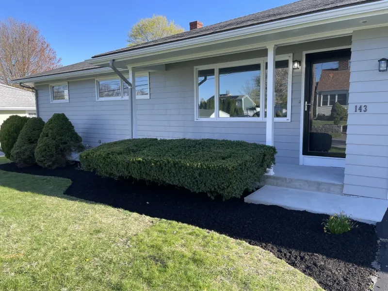 Light gray house with green shrubs, black mulch, and a glass front door on a sunny day.