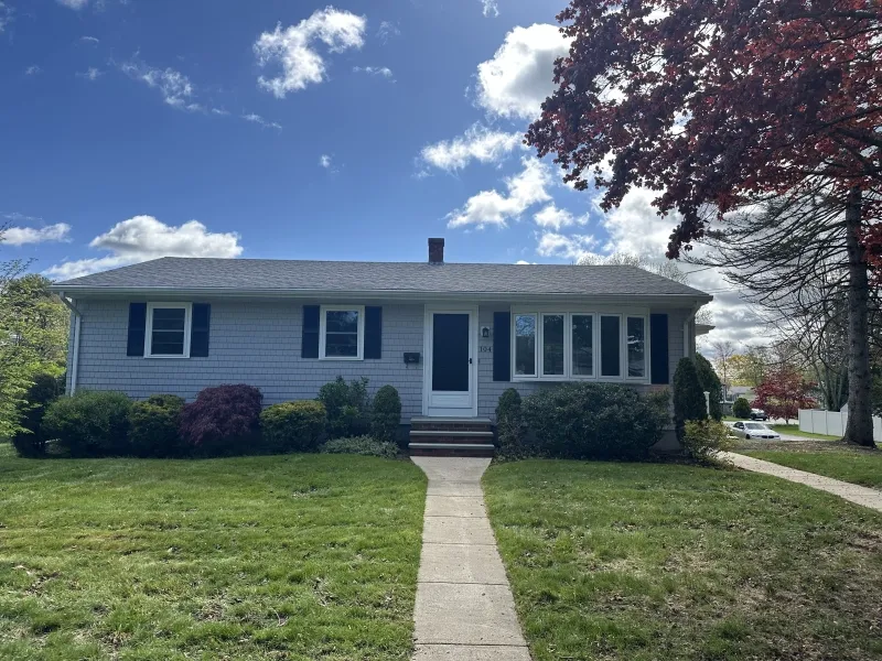 Single-story gray house with a front lawn, sidewalk, and trees under a partly cloudy sky.