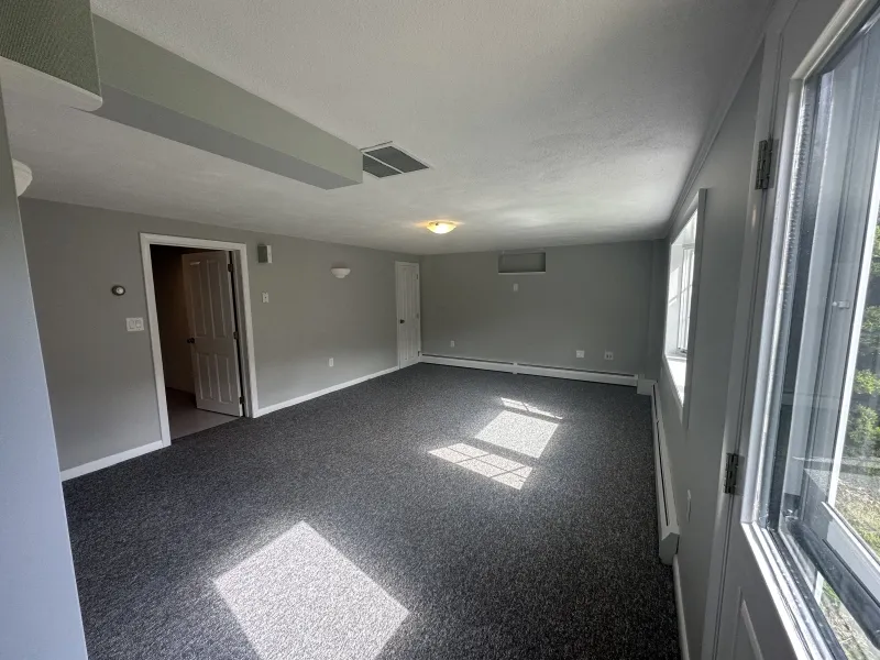 A spacious, empty gray-carpeted room with gray walls and sunlight coming through windows.