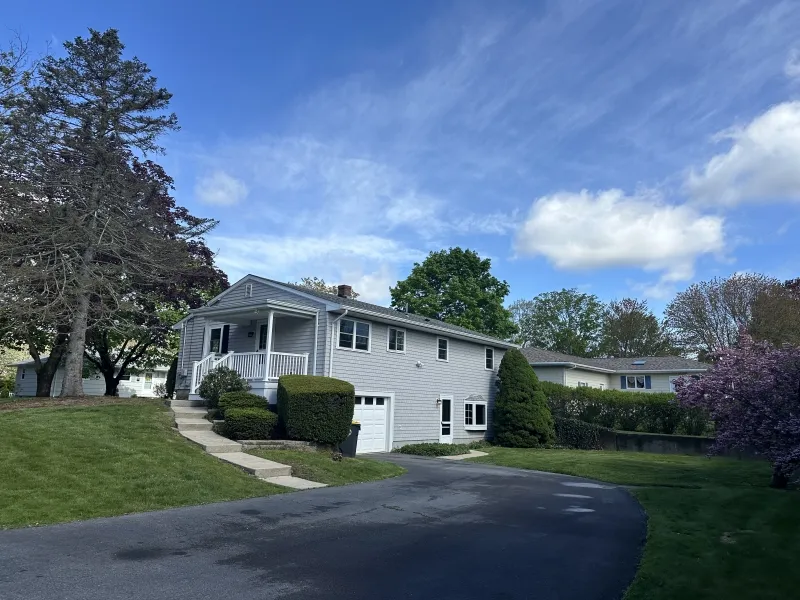 Gray two-story house with porch, driveway, and green lawn under a blue sky with clouds.