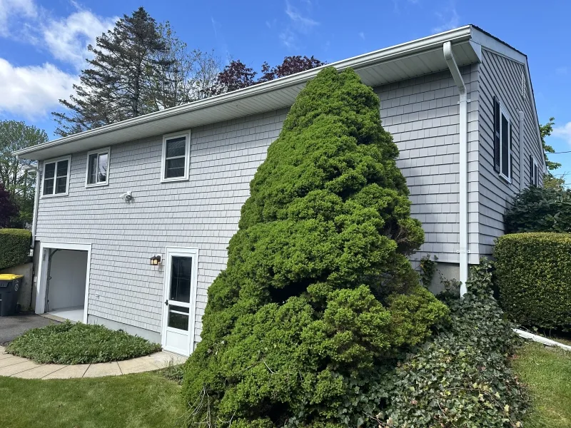 Gray sided house with upper windows and a large green bush in front, under a blue sky.
