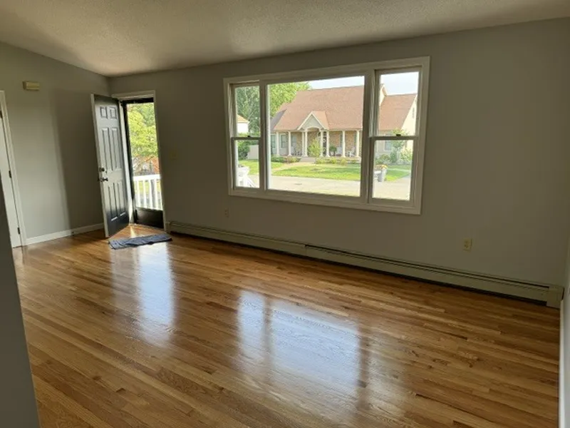 Bright, empty living room with wood floors and large window facing a house across the street.
