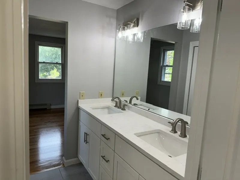 Modern bathroom with double sinks, large mirror, and bright lighting, view into another room.