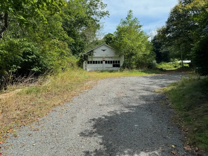 A gravel driveway leads to a white garage surrounded by green trees and overgrown grass.