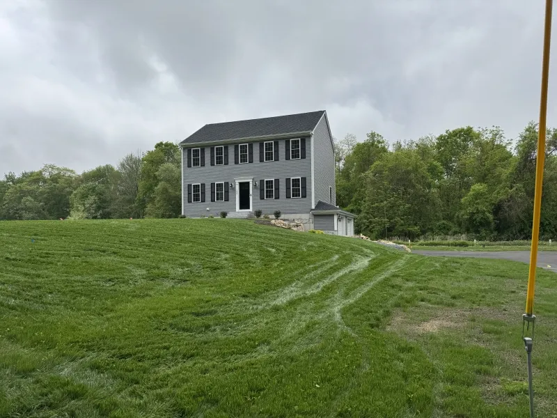 Gray two-story house on a grassy hill with cloudy skies and trees in the background.