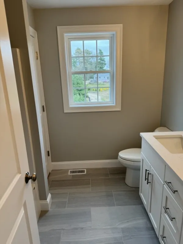Small bathroom with gray tile floor, white vanity, toilet, and a window with outdoor view.