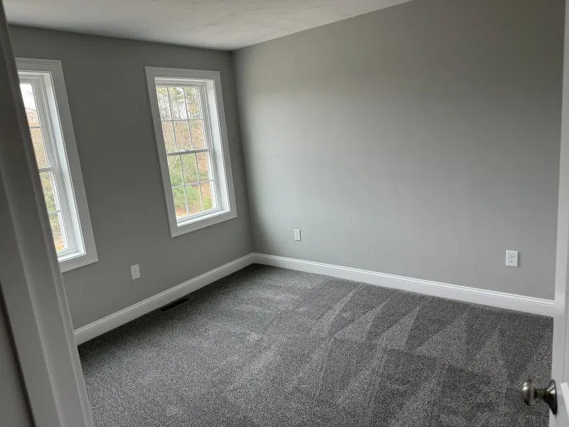 Empty room with gray carpet, light gray walls, and two large windows letting in natural light.