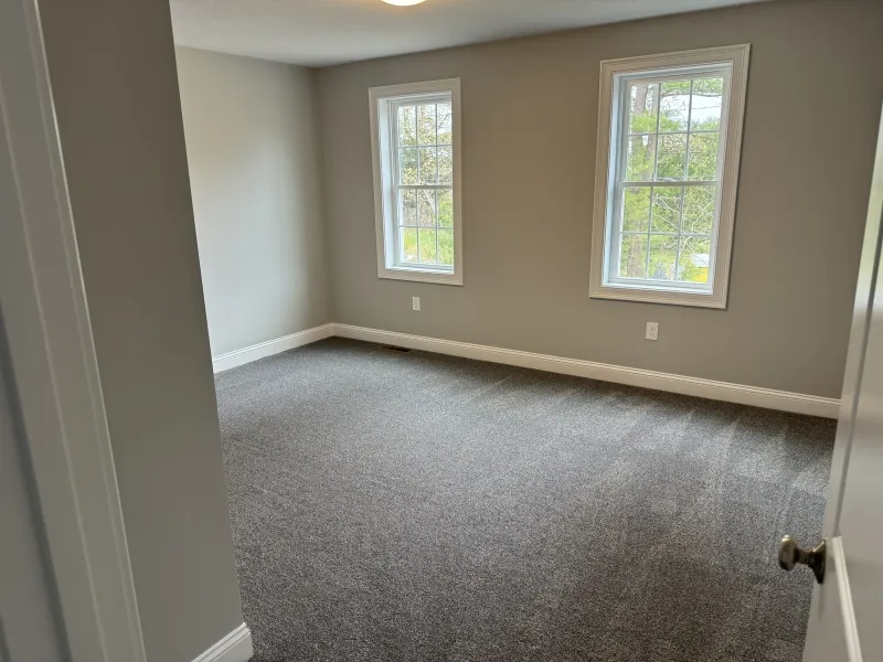 Empty room with gray carpet, light gray walls, and two windows letting in natural light.
