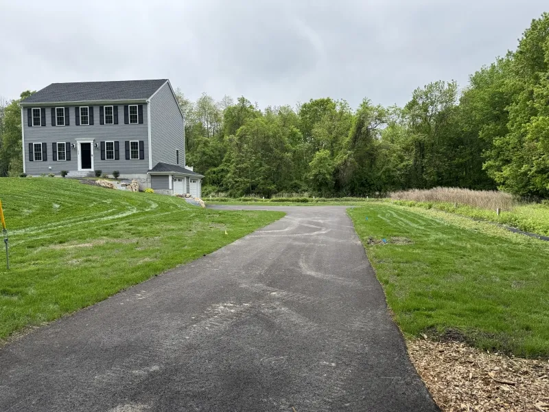 A paved driveway leads to a gray two-story house with a green lawn and trees behind.