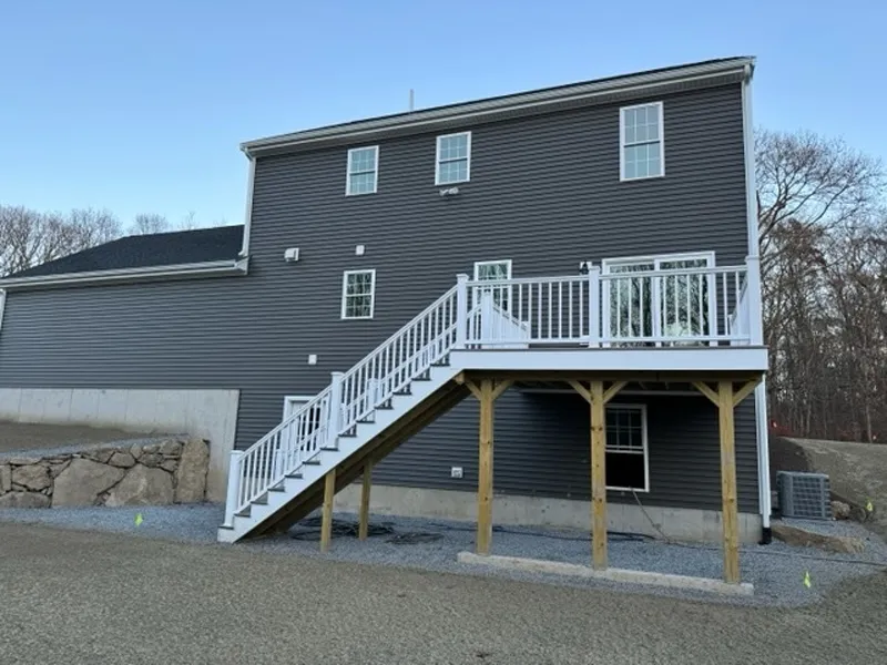 Gray house with white-railed deck and stairs, supported by wooden posts, gravel ground.