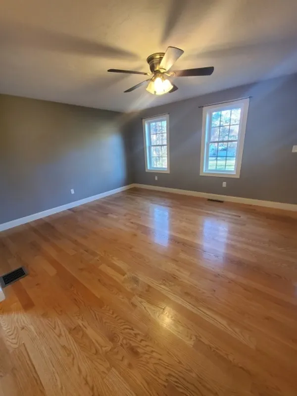 Empty room with wood floors, gray walls, two windows, and a ceiling fan with lights.
