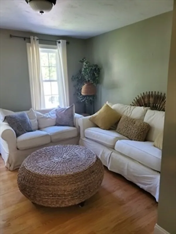 Cozy living room with white sofas, woven ottoman, pillows, plant, and sunlight through window.