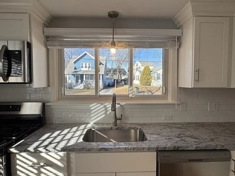 A kitchen sink under a window with sunlight streaming in, showing houses outside.