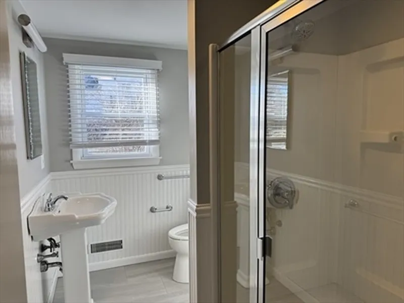 Small bathroom with a pedestal sink, toilet, window, and glass shower enclosure.