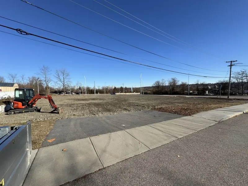 A small excavator sits on a cleared, empty lot next to a sidewalk under a clear sky.