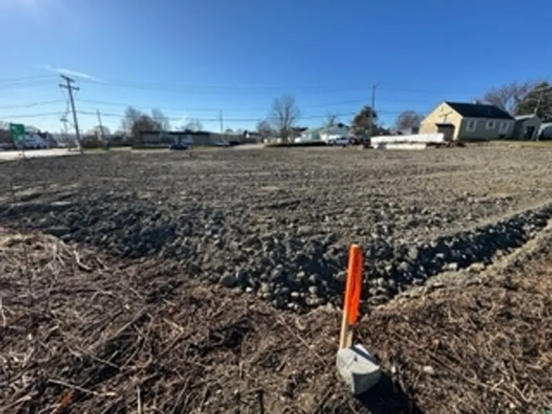 A cleared, empty lot with a survey marker and houses in the background under a blue sky.