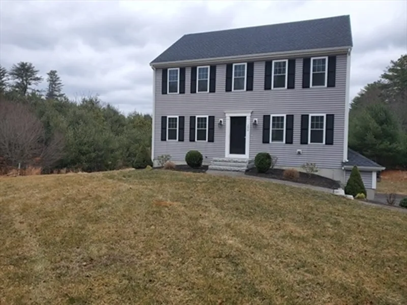 Two-story gray house with black shutters, surrounded by grass and trees on a cloudy day.