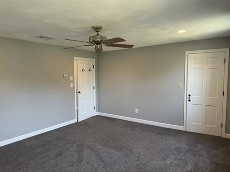 Empty room with gray walls, ceiling fan, two white doors, and dark carpeted floor.