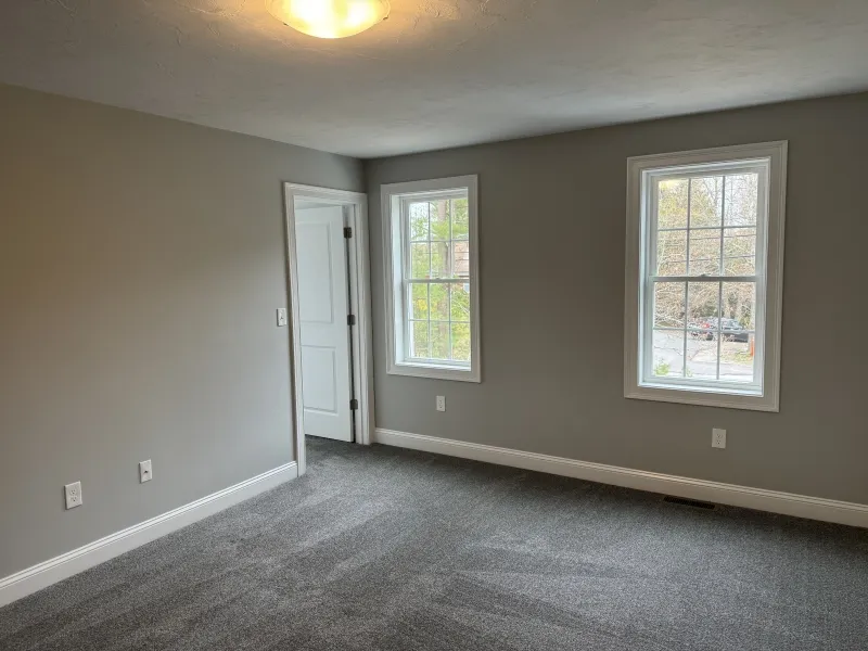 Empty room with gray carpet, light gray walls, two windows, and a white door.
