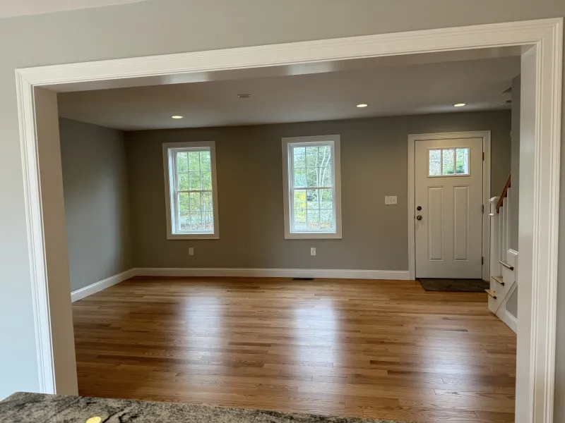 Empty living room with hardwood floors, gray walls, two windows, and a white front door.