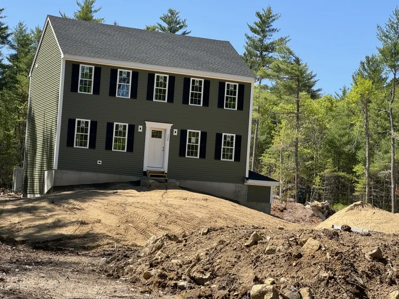 A green house on a sloped, unfinished dirt lot surrounded by trees.