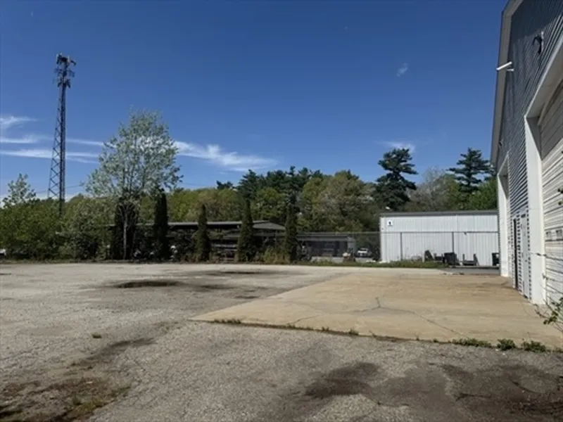 Empty paved lot beside a white industrial building, with trees and blue sky in the background.