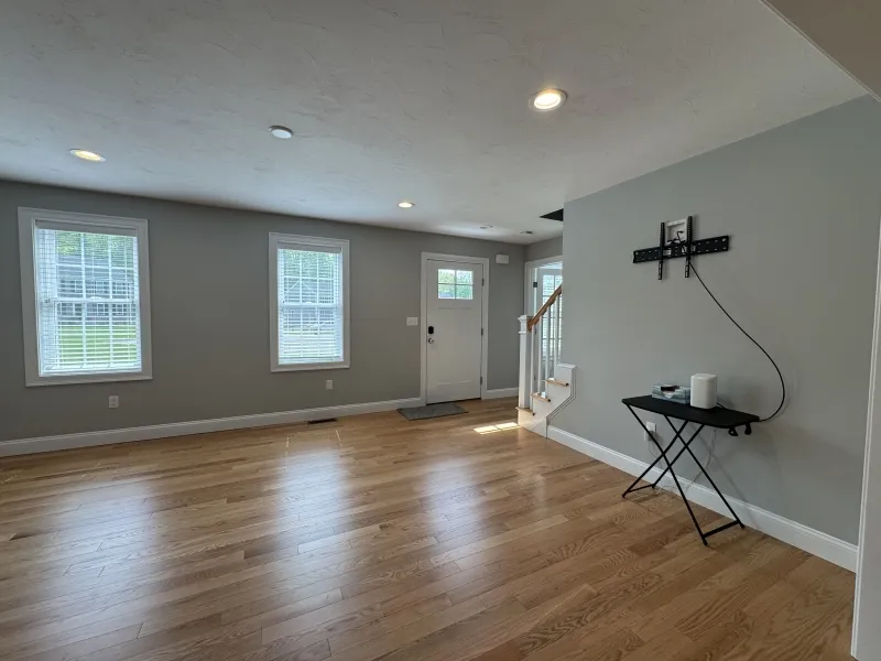 Bright, empty living room with wood floors, gray walls, and three windows by the front door.