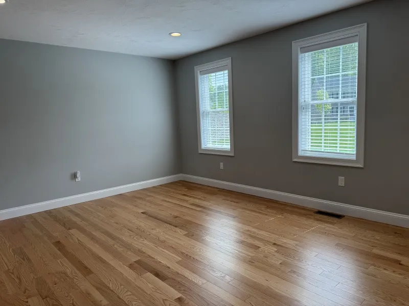 Empty room with light hardwood floor, gray walls, and two windows with white blinds.