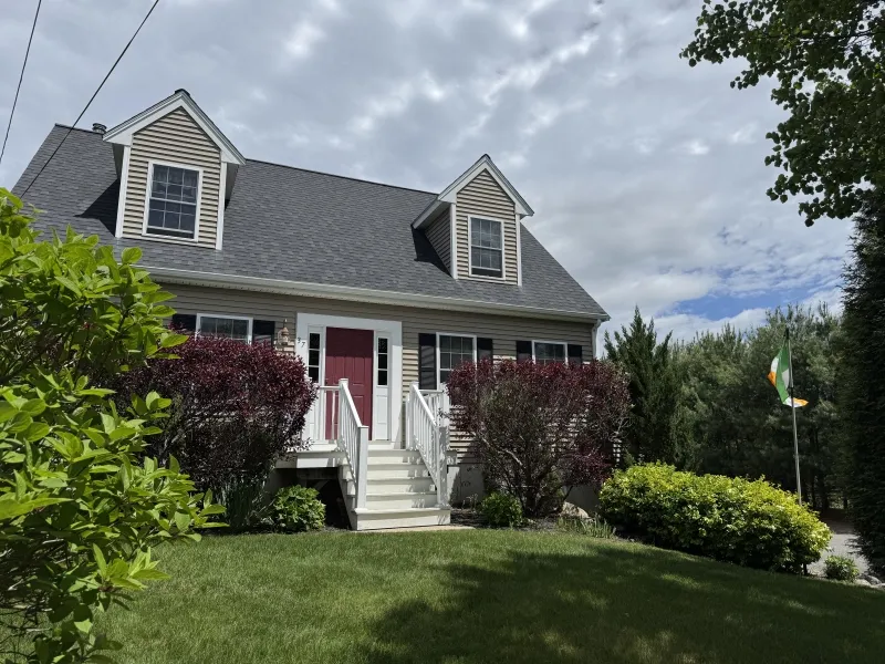 A two-story house with a red door, front steps, shrubs, and a green lawn under a cloudy sky.