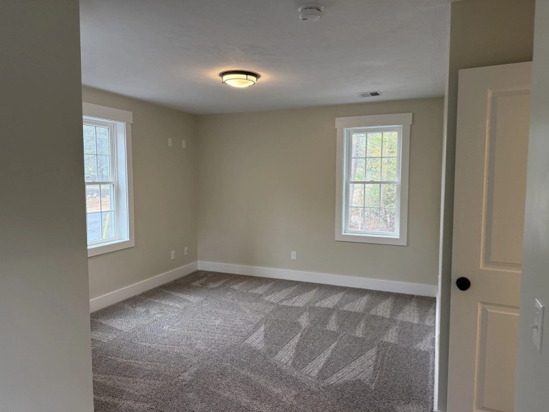 Empty room with beige walls, gray carpet, two windows, and an open white door.