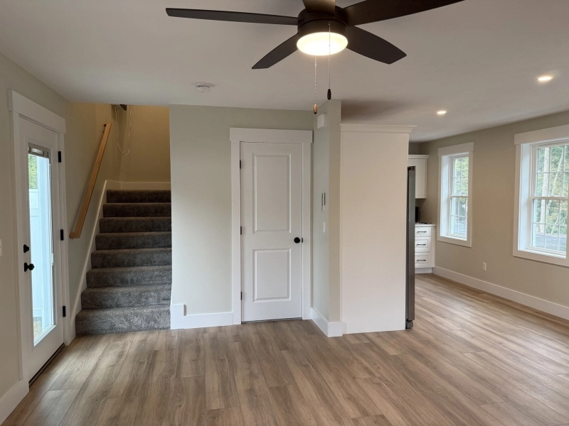 Bright room with wood floors, ceiling fan, stairs, white door, and large windows.