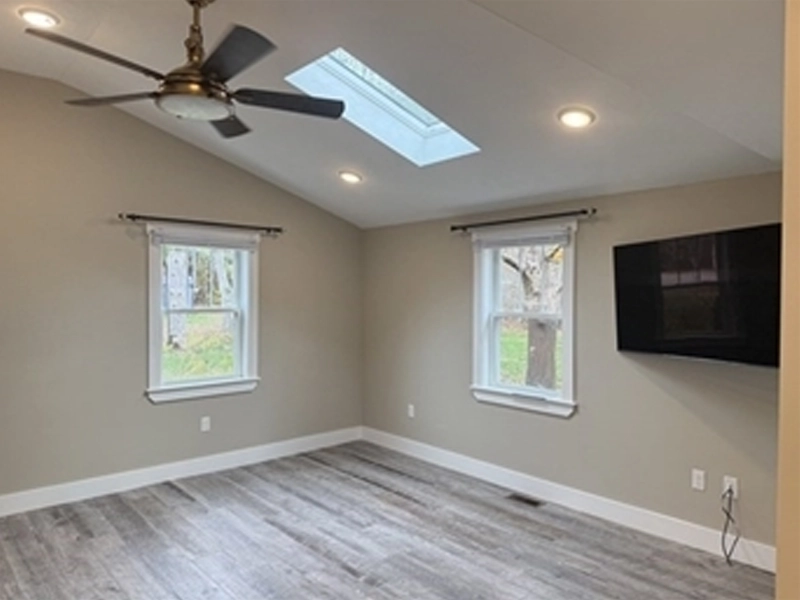 Empty room with wood floors, ceiling fan, skylight, two windows, and a wall-mounted TV.